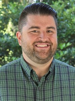 Man in green plaid button down shirt with styled hair smiling at camera in front of a tree